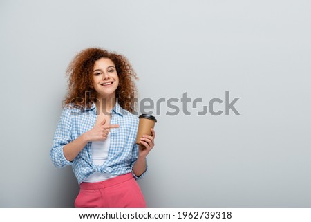 pleased woman pointing at paper cup while looking at camera on grey background