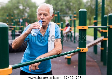 mature peppy pensioner man resting and drinking water on a sports equipped playground