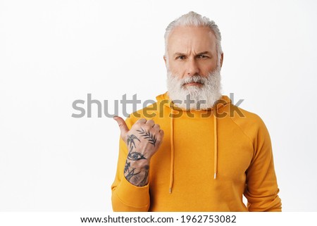 Disappointed serious old man with tattoos, pointing left and looking angry at camera, asking question about logo brand, need explanation, scolding someone, white background