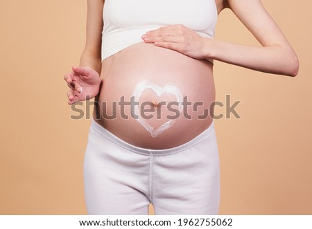 Close-up, a pregnant girl, in a white sports top and leggings, holds her hands on her bare belly with heart-shaped cream on it. Concept for motherhood, childbirth, stretch marks, pregnant skin care.