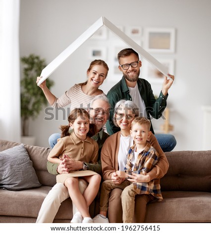 Big happy family, grandparents, mother, father with little kids son and daughter celebrating relocation in new home, sitting on the sofa under paper roof and smiling at camera. Mortgage loan concept Royalty-Free Stock Photo #1962756415