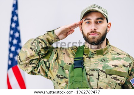 of handsome soldier in a military uniform salutes and looks to the camera near the American flag in studio on white background
