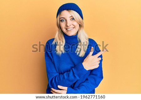 Young caucasian woman wearing wool winter sweater and cap smiling cheerful pointing with hand and finger up to the side 