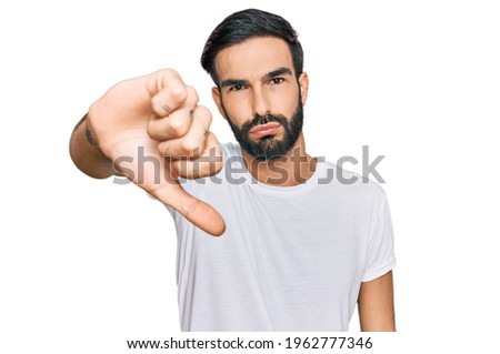 Young hispanic man wearing casual white t shirt looking unhappy and angry showing rejection and negative with thumbs down gesture. bad expression. 