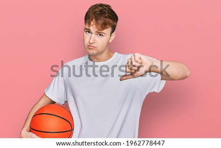 Young caucasian man holding basketball ball with angry face, negative sign showing dislike with thumbs down, rejection concept 