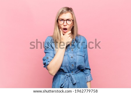 young blond pretty woman with mouth and eyes wide open and hand on chin, feeling unpleasantly shocked, saying what or wow