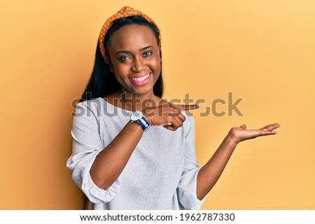 Young african woman wearing casual clothes over yellow background amazed and smiling to the camera while presenting with hand and pointing with finger. 