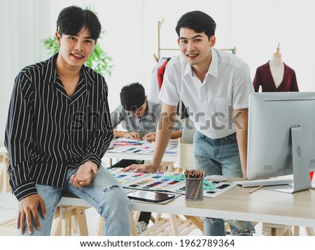 Asian dressmaker using tablet near male colleagues browsing data on computer and drawing sketch in atelier