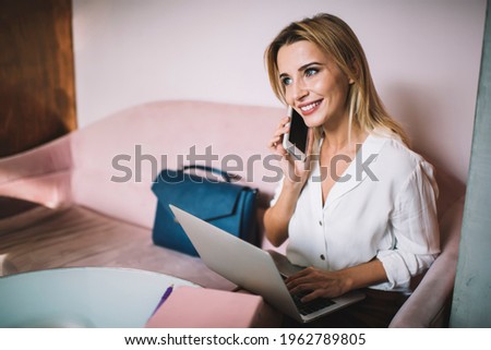 Young female entrepreneur in formal clothes sitting at table with laptop and talking on phone while working on project in modern workspace