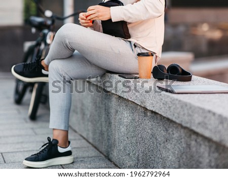 Study and work remotely online outdoors during COVID-19 lockdown. Young male hipster worker in stylish clothes sits on street with gadgets, headphones and cup of takeaway coffee, next to bike, cropped