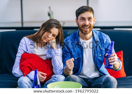 Bored wife annoyed by horny husband, soccer fan celebrating victory in sports game with beer in hand, sitting on the couch at home.