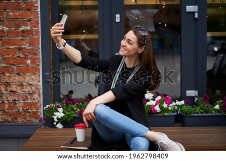 Happy Caucaisan woman clicking selfie images during leisure time using mobile application outdoors, cheerful hipster girl in casual clothes smiling at front smartphone camera during leisure in city