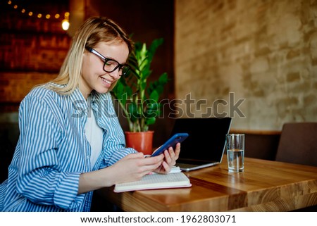 Millennial female student in classic spectacles browsing web publication on cellphone device using coworking internet connection while e learning, happy Caucasian blogger enjoying network chatting