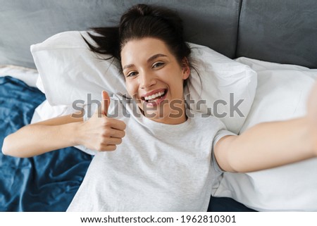 Brunette woman showing thumb up and taking selfie photo on bed at home
