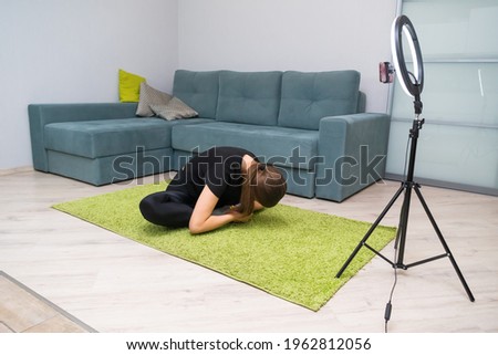 Young beautiful dark-haired female blogger writes content for her video blog. Attractive woman is engaged in fitness, stretching at home on a light green mat. Concept of fitness and healthy lifestyle