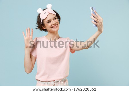 Young happy woman in pajamas jam sleep eye mask rest relaxing at home doing selfie shot on mobile phone waving hand isolated on pastel blue background studio portrait. Good mood night bedtime concept