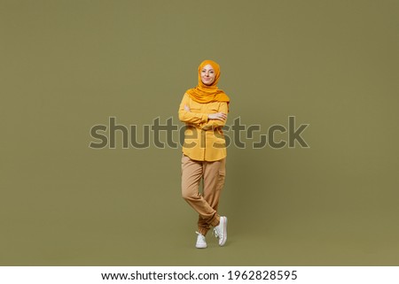 Full length young arabian asian muslim woman in abaya hijab yellow clothes hold hands crossed folded isolated on olive green khaki background studio. People uae middle eastern islam religious concept Royalty-Free Stock Photo #1962828595