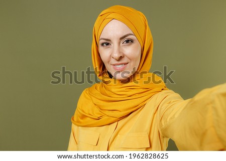Close up young smiling happy arabian asian muslim woman 20s in abaya hijab yellow clothes doing selfie shot on mobile phone isolated on olive green background studio People uae islam religious concept