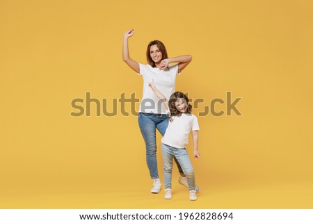 Full length happy woman in basic white t-shirt have fun with cute child baby girl 5-6 years old hold hands. Mom little kid daughter isolated on yellow color background studio. Mother's Day love family