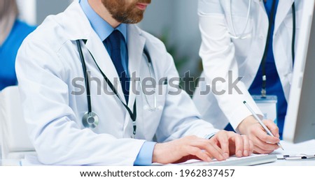 Close up of mixed-race doctors speaking in hospital cabinet at work. Caucasian young handsome male physician typing on computer and talking with Asian female colleague. Healthcare, clinic concept