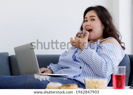 Asian young happy beautiful fat female shopper sit relaxing on fabric sofa look at camera using laptop computer searching products shopping online while eating delicious chocolate donut in hand. Royalty-Free Stock Photo #1962847903