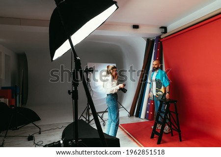 Woman photographer with a camera in her hands in a photo studio makes a photo shoot for a handsome bearded man on a red background.