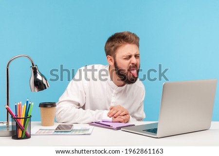 Funny displeased man with beard showing tongue out talking video call on laptop sitting at workplace, having fun, demonstrating childish behavior. Indoor studio shot isolated on blue background Royalty-Free Stock Photo #1962861163