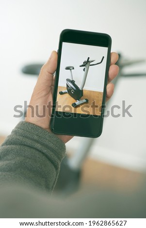closeup of a young caucasian man taking a picture with his smartphone of his used stationary bicycle, to sell it or exchange it on an online marketplace app to sell and buy secondhand goods