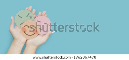 Woman hand holding emotion group of paper cut happy smile face, angry and boring face on blue background, positive and negative thinking, mental health assessment , world mental health day concept