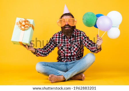 funny brutal bearded man wear checkered shirt having lush beard and moustache hold party balloons and present box, birthday