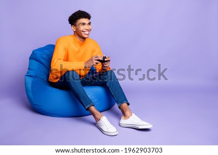 Full length body size photo of guy sitting in beanbag looking copyspace playing video games with joystick isolated pastel violet color background