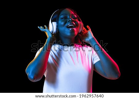 Enjoying music, dancing. African young woman's portrait on dark studio background in neon light. Beautiful female model in headphones. Concept of human emotions, facial expression, youth, sales, ad.