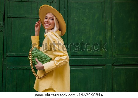 Joyful smiling woman wearing yellow hat, classic shirt, holding quilted faux leather green bag with chain, posing on green background in street. Copy, empty space for text Royalty-Free Stock Photo #1962960814