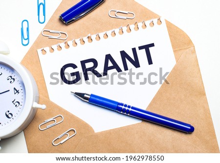 On a light background, a kraft envelope, an alarm clock, paper clips, a blue pen and a sheet of paper with the text GRANT.