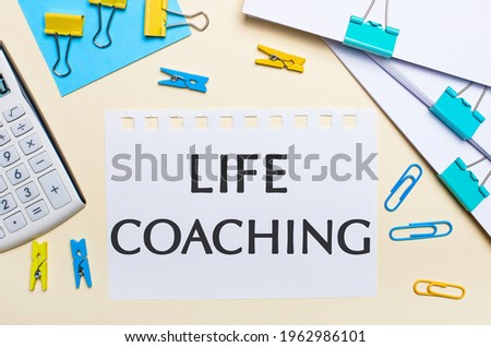 On a light background, there are stacks of documents, a white calculator, yellow and blue paper clips and clothespins, and a notebook with the text LIFE COACHING