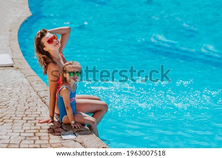 Young pretty woman and cute little girl in summer bikini and sunglasses posing together sitting near the pool in hotel. Copy space.