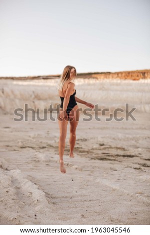 Beautiful blonde sporty girl in a black bodysuit runs on sand quarry on sunny day.