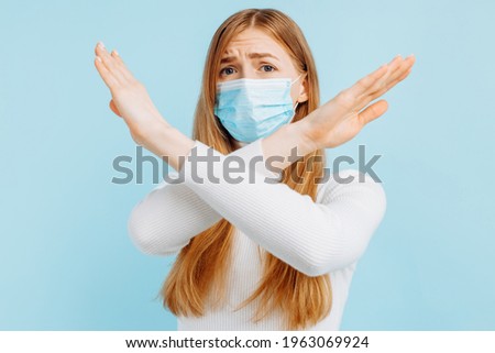 Young woman in a medical protective mask to protect against viruses, crossing her arms, shows a stop gesture to stop action, on a blue background
