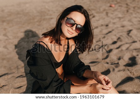 Adorable pretty woman with dark hair and black shirt posing at camera while resting on the beach in sunlight. High quality photo