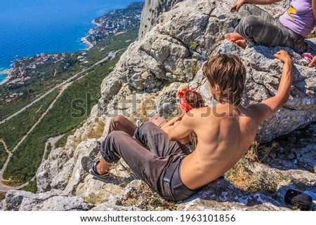 The girls and the guy are sitting on the edge of the mountain. Below you can see the blue sea. Girl photographing. Crimea
