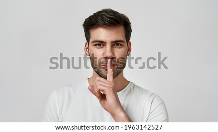 Silence gesture. Young bearded man in white t-shirt keeping finger on his lips while standing against grey background. Studio photo concept