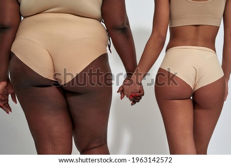 Cropped shot of two african american women in underwear with different body size holding each other hands, standing together isolated over gray background. Diversity, body positive concept. Back view