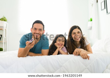 Happy family watching a comedy movie on the tv while lying in bed. Mom, dad and girl laughing and enjoying cartoons during a lazy morning in the bedroom