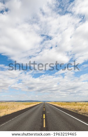 A straight road of blacktop going to a vanishing point in the open and flat country of Grant County in Eastern Washington State.  The cloud layer above is broken with pockets of blue sky Royalty-Free Stock Photo #1963219465