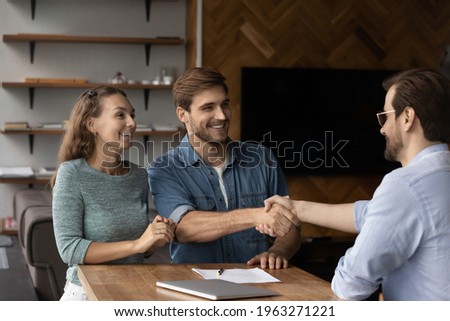 Smiling young Caucasian couple shake hand with male realtor or broker at office meeting. Happy man and woman clients handshake close deal make agreement with real estate agent. Rental, rent concept. Royalty-Free Stock Photo #1963271221