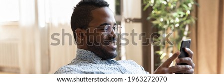 Happy millennial guy using online bank app on smartphone for payment. African man reading article on internet on cell phone, ordering on store website, making video call from home. Panorama photo