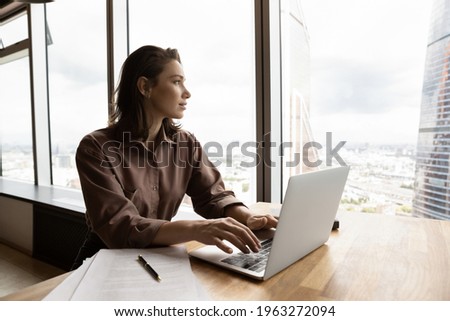 Dreamy young Caucasian businesswoman work on computer in modern office building look in distance making plans of career success or perspectives. Pensive female employee busy with laptop thinking. Royalty-Free Stock Photo #1963272094