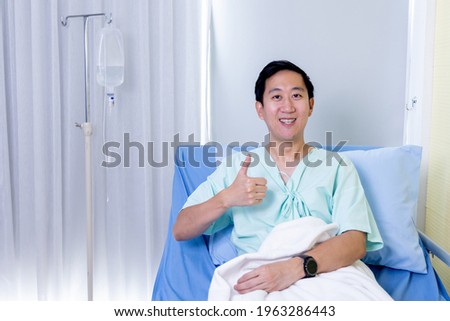 Portrait of a smiling Asian young male patient sitting on hospital bed room showing thumbs up gesture and sign to camera