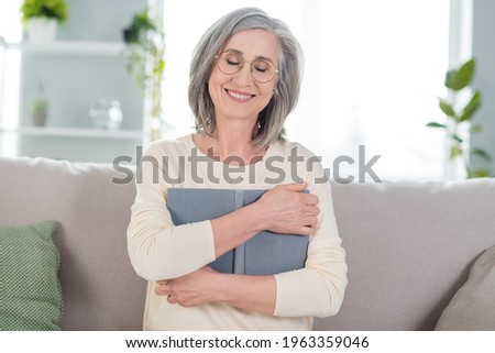 Photo of peaceful happy charming old woman hug embrace book reader sit couch indoors inside house home