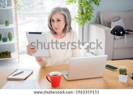 Photo of calm serious old woman look use tablet wear glasses sit desk focused indoors inside house home